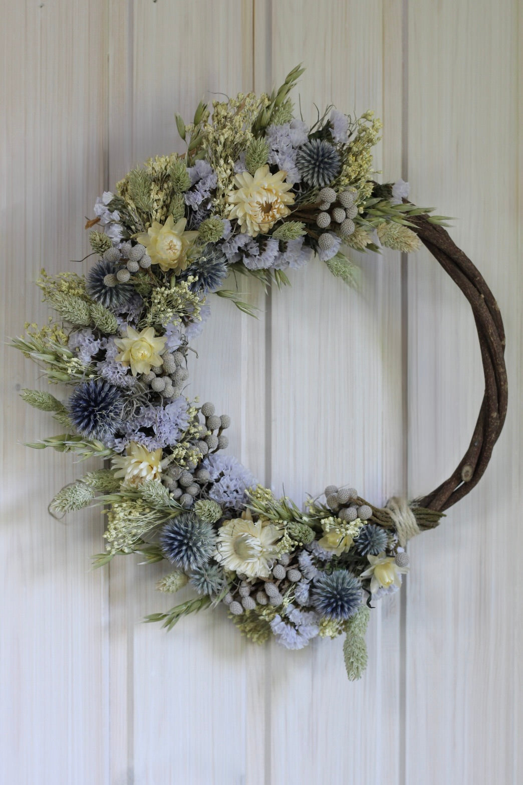 Handmade dried flower wreath in soft lavender, cream, and grey-blue tones on a 25cm twisted vine base, featuring strawflowers, globe thistles, statice, and natural grasses.