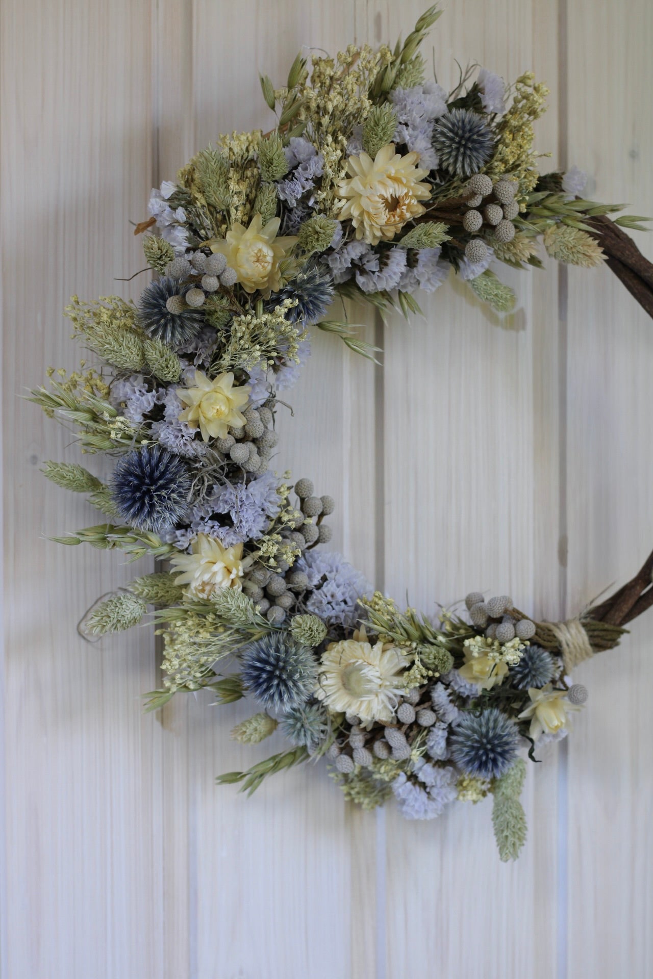 Handmade dried flower wreath in soft lavender, cream, and grey-blue tones on a 25cm twisted vine base, featuring strawflowers, globe thistles, statice, and natural grasses.