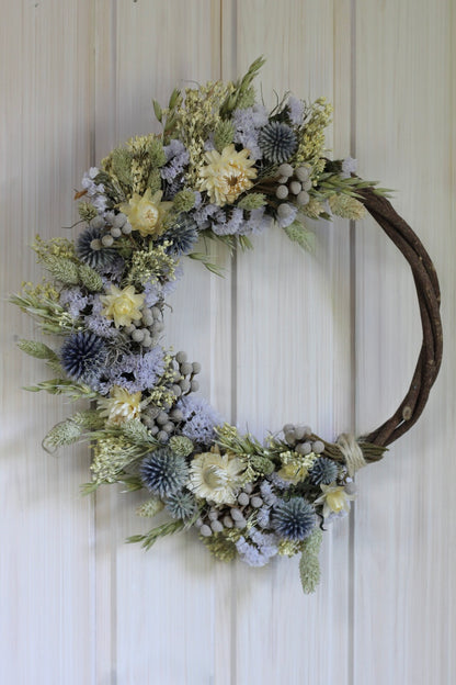 Handmade dried flower wreath in soft lavender, cream, and grey-blue tones on a 25cm twisted vine base, featuring strawflowers, globe thistles, statice, and natural grasses.