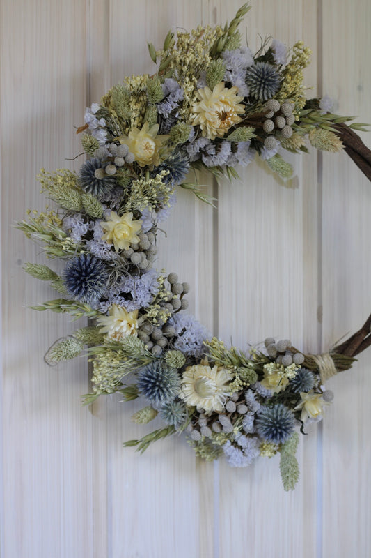Handmade dried flower wreath in soft lavender, cream, and grey-blue tones on a 25cm twisted vine base, featuring strawflowers, globe thistles, statice, and natural grasses.